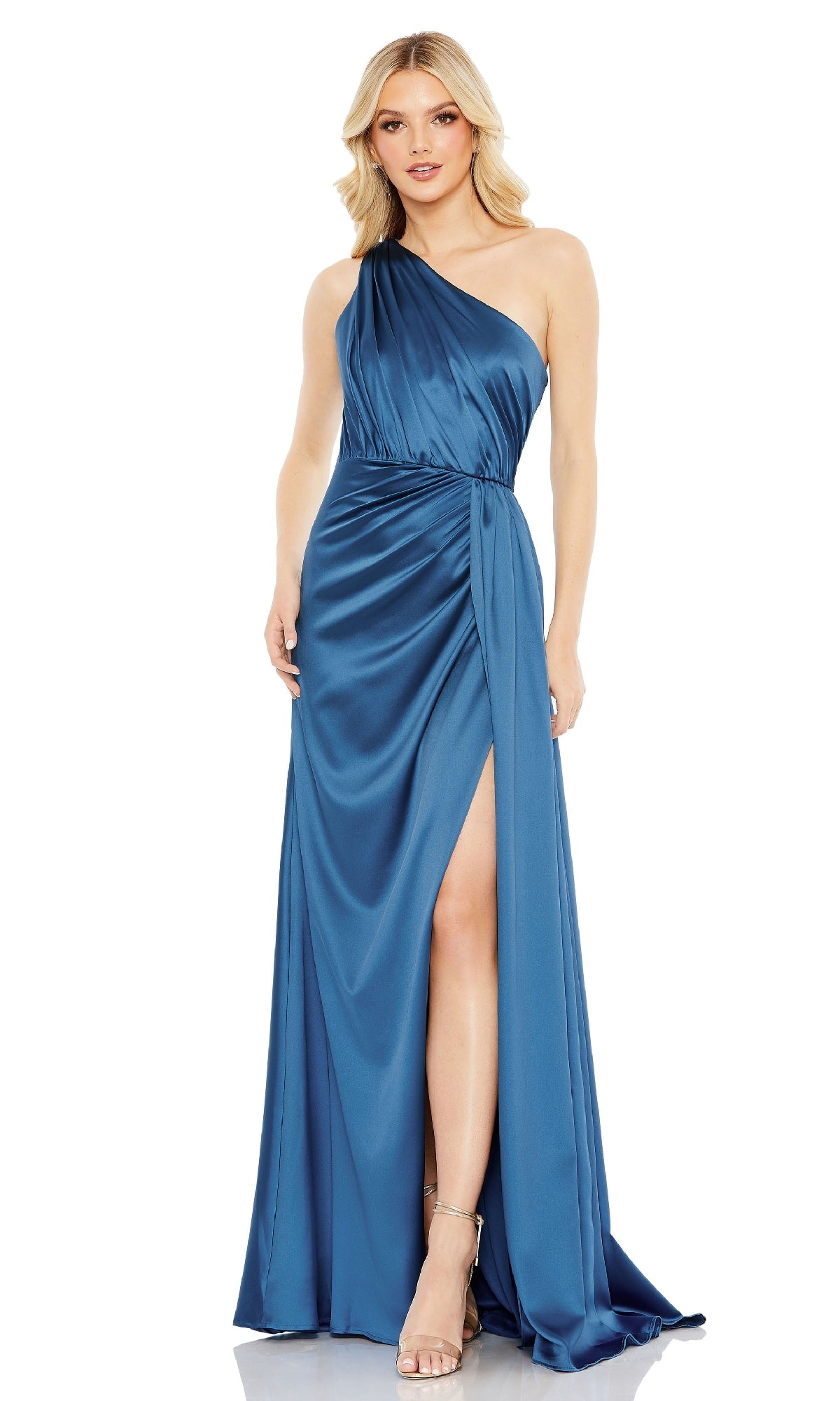 A woman stands against a white background wearing the Mac Duggal 26654 one-shoulder long formal dress in blue satin with a thigh-high slit and silver high-heeled sandals.